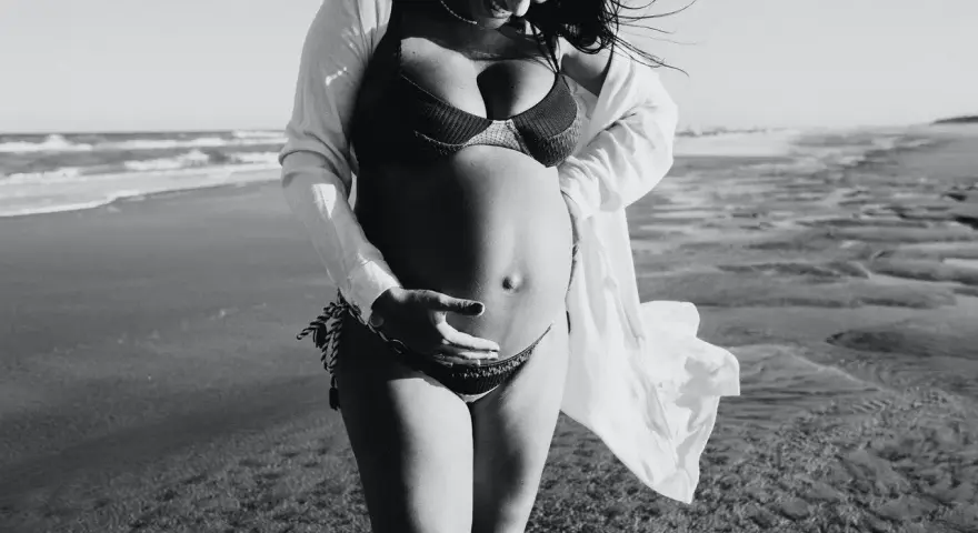 Donna incinta si gode il sole in spiaggia, accarezzando il pancione. La brezza marina le scompiglia i capelli.