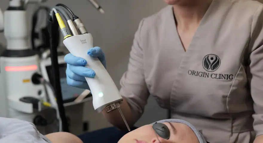 Zabieg laserem Fotona R35NX w Origin Clinic. Widoczna głowica lasera i pacjentka w okularach ochronnych. Oczekiwane spektakularne efekty odmładzające.