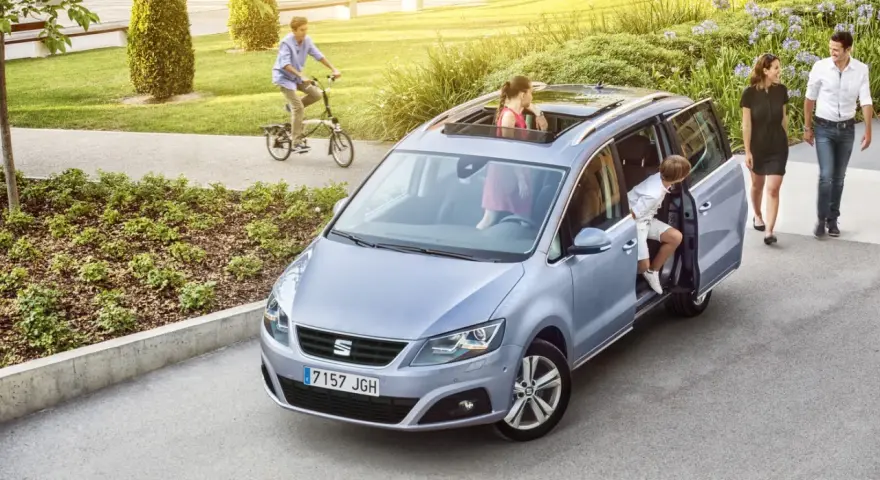 Familienauto mit zwei Kindern, die ein- und aussteigen. Ein Junge fährt Fahrrad im Hintergrund.