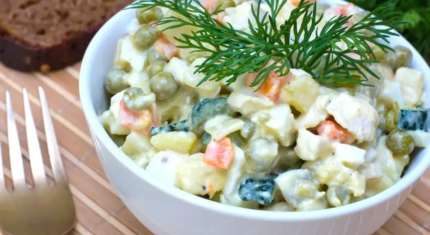 Ein Schüsselchen Olivier Salat, garniert mit Dill, neben einer Scheibe dunklem Brot und einer Gabel.