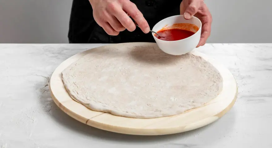 Przygotowanie ciasta na pizzę bez drożdży. Dłonie nabierają sos pomidorowy z miseczki, by rozprowadzić go na okrągłym spodzie.