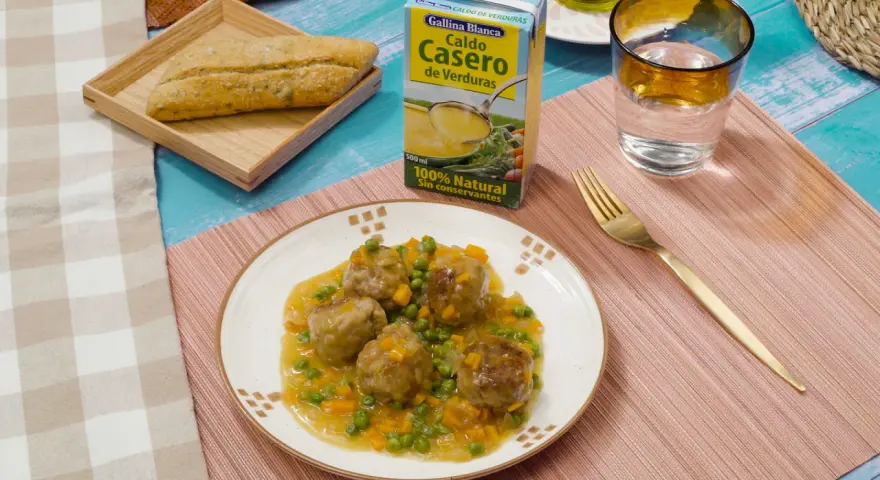 Albondigas a la jardinera en un plato blanco, acompañadas de un pan rústico, un vaso de agua y un caldo de verduras.