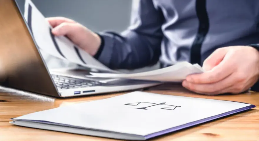 Osoba przegląda dokumenty przy laptopie. Na pierwszym planie folder z symbolem wagi, sugerujący sprawy związane z prawem i potencjalną masą upadłościową.
