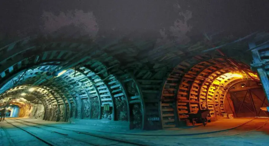 Wnętrze kopalni Nowa Ruda. Tunel oświetlony niebieskim i pomarańczowym światłem, z drewnianymi podporami i torami kolejowymi.
