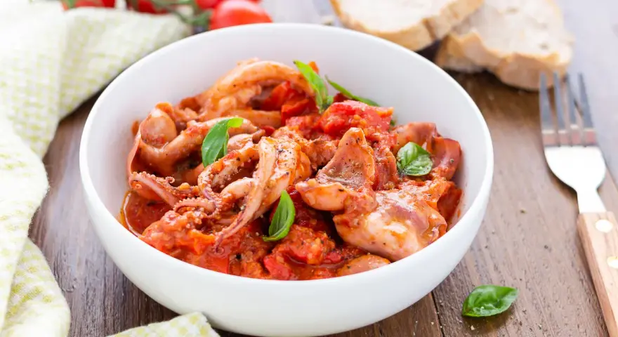 Totani in umido con pomodoro e basilico, serviti in una ciotola bianca, con pane e forchetta sullo sfondo.