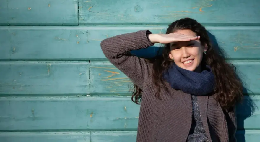 Ragazza sorride, coprendosi gli occhi dal sole. Il fastidio alla luce non le impedisce di godersi la giornata.