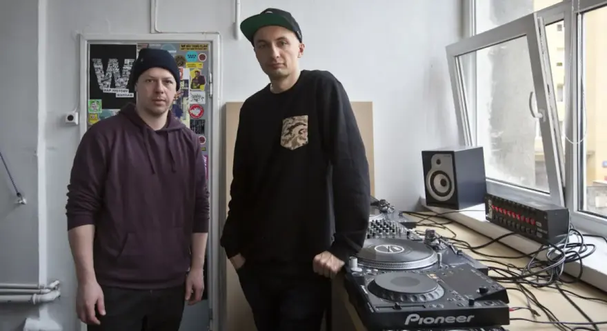 Dwóch mężczyzn przy sprzęcie DJ-skim. Jeden w bordowej bluzie z kapturem, drugi w czarnej bluzie z naszywką. To ich sposób na pokazanie, co to stereo.