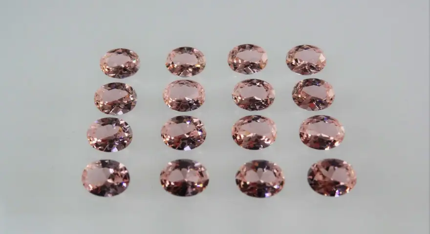 Sedici pietre di morganite ovali, rosa pesca, disposte in file ordinate. Le loro sfaccettature riflettono la luce, mostrando le belle morganite proprietà.