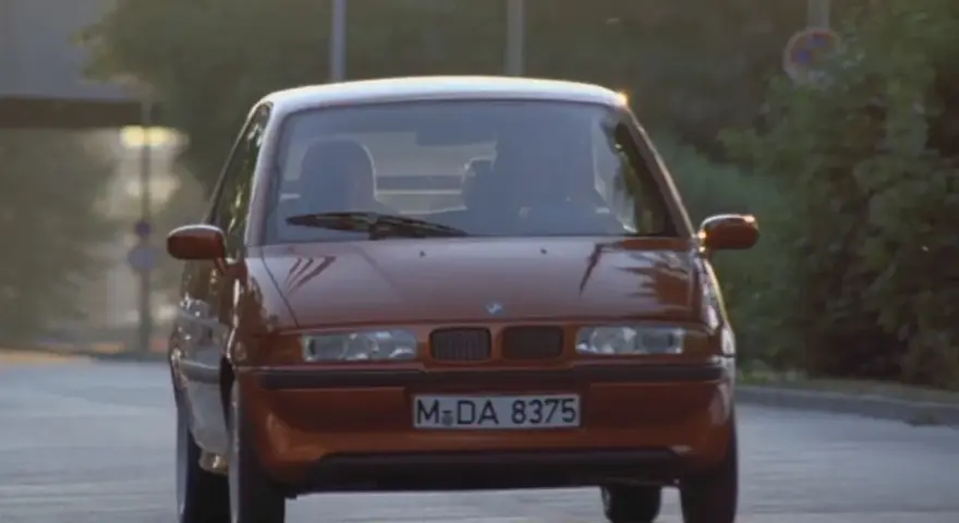 Pomarańczowy BMW E1 jedzie po drodze, światła odbijają się od karoserii.