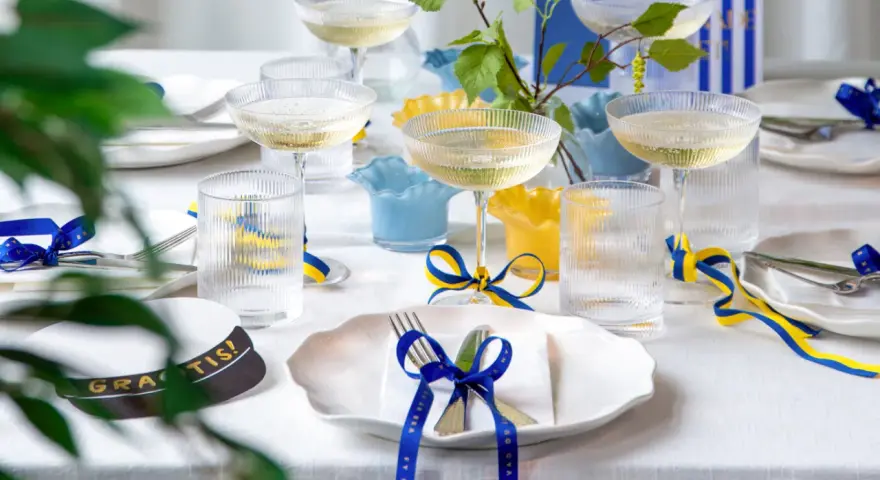 Festligt dukat bord med champagneglas, blå och gula band som knyts i en blå rosett, samt en studentmössa.