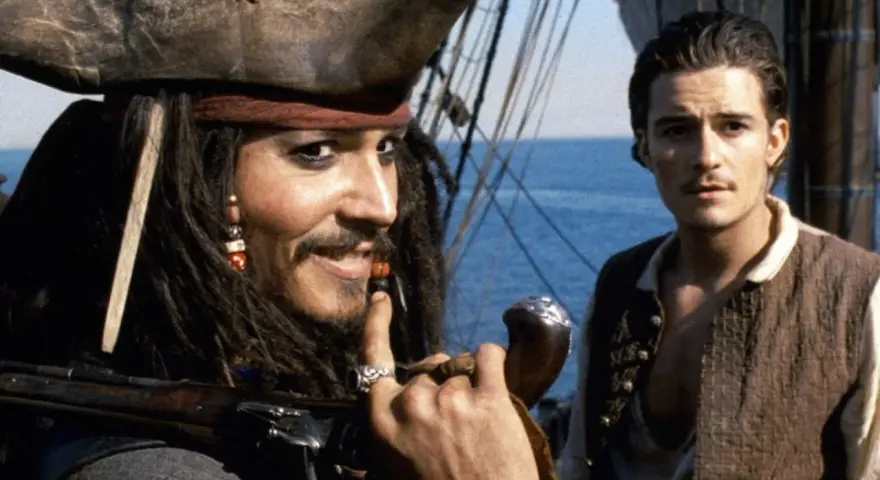 Jack Sparrow, z fajką i karabinem, uśmiecha się do Willa Turnera. Pewnie myśli o kolejnej butelce rumu.