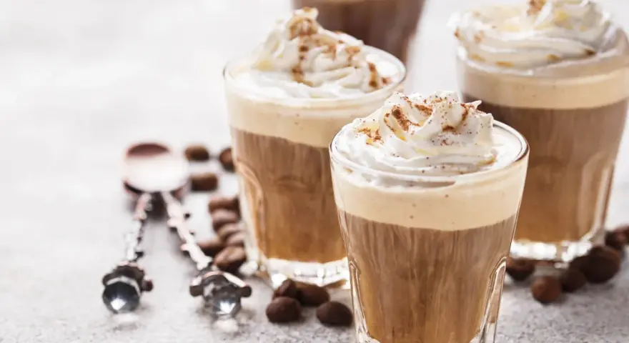 Tre bicchieri di deliziosa crema al caffè, guarniti con panna montata e cacao. Chicchi di caffè e cucchiaini decorati completano la scena.