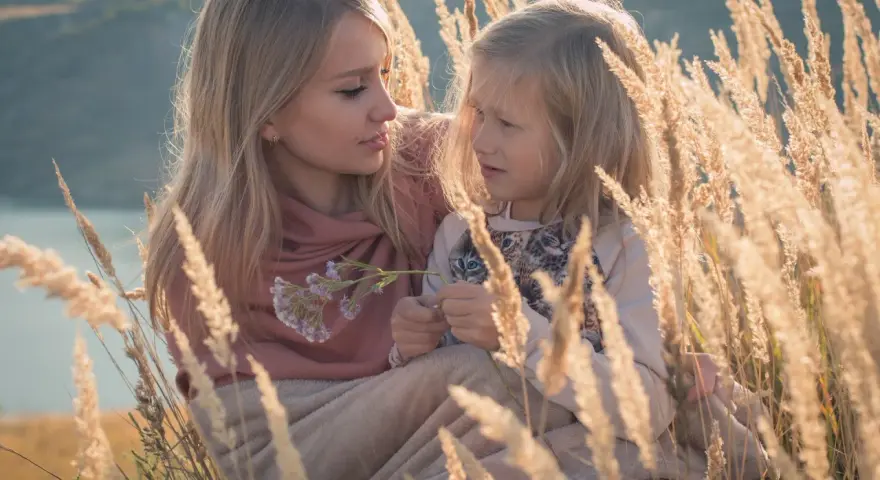 Matka i córka w trawach. Córka z autyzmem skupiona na kwiatku, matka z czułością patrzy na nią.