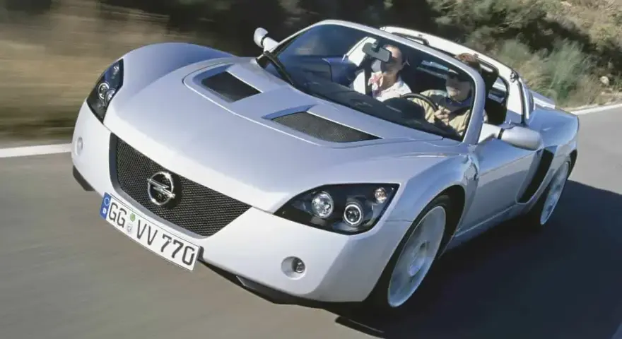 Srebrny Opel Speedster z dwoma pasażerami pędzi po krętej drodze.