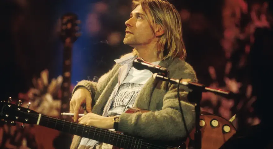 Kurt Cobain z papierosem w dłoni, grający na gitarze. Jego muzyka to część 100 najlepszych piosenek wszechczasów.
