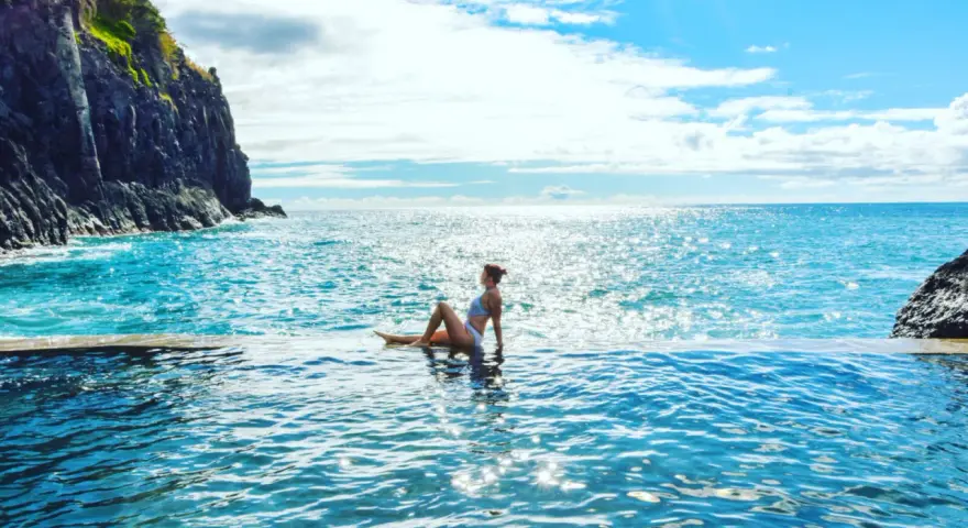 Madera, listopad. Słoneczna pogoda zachęca do kąpieli w basenie z widokiem na ocean. Kobieta w białym bikini relaksuje się na brzegu.