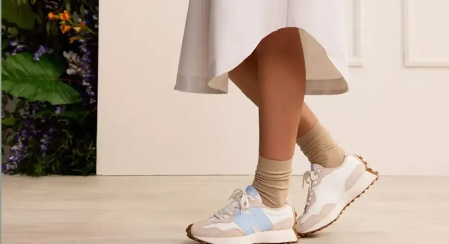 Elegancka sukienka i sportowe buty tworzą stylowy kontrast. Nogi w beżowych skarpetkach i białych sneakersach z błękitnym logo.