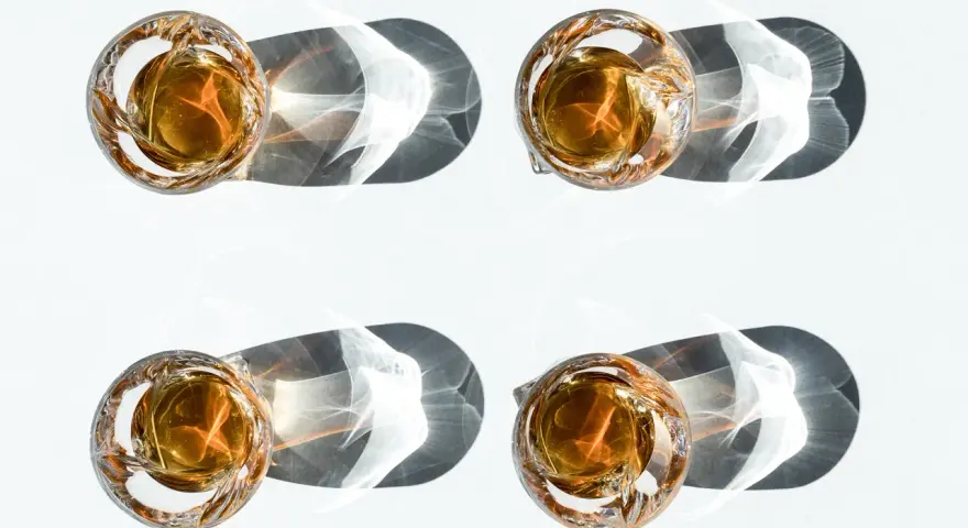 Vier Gläser mit bernsteinfarbenem Whisky werfen dramatische Schatten. Ein visueller Vergleich, der den Unterschied Scotch und Bourbon verdeutlicht.