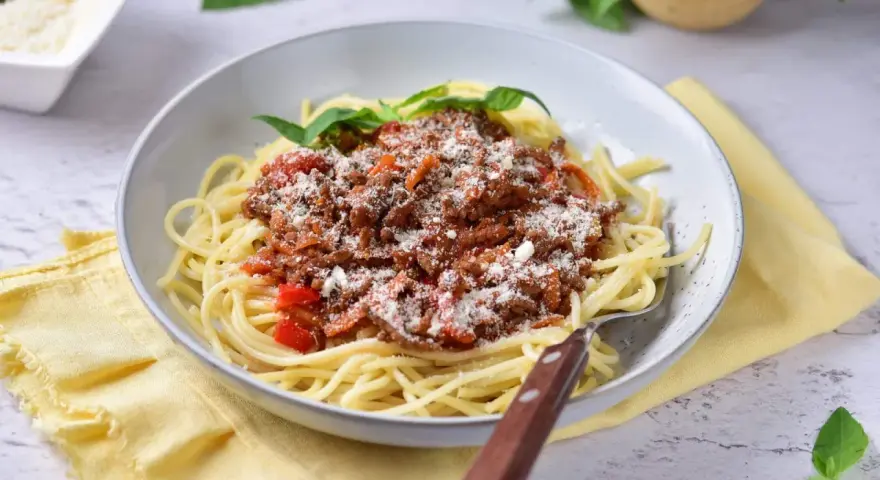 Pyszne spaghetti z bogatym sosem mięsnym na bazie passaty, posypane parmezanem i udekorowane listkami bazylii.