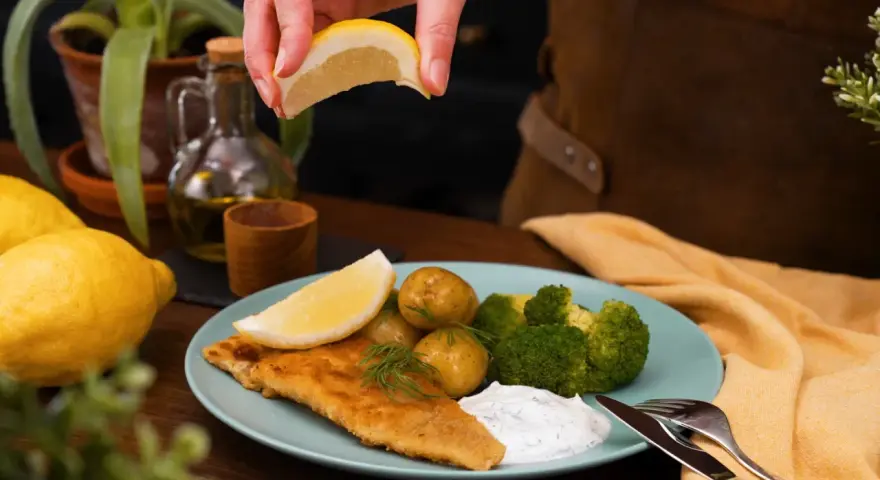 En hand pressar citron över en tallrik med panerad torsk, potatis, broccoli och en klick sås.