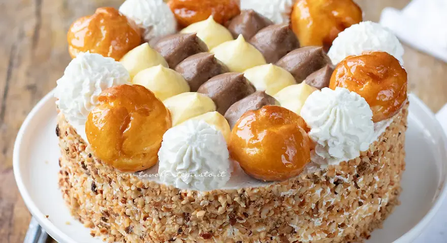 Una deliziosa torta Saint Honoré, con bignè caramellati, panna montata e crema al cioccolato e vaniglia, il tutto ricoperto di granella di nocciole.