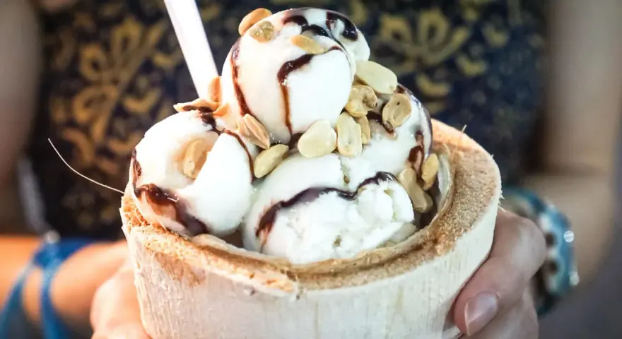 Cremiges Pina Colada Eis in einer Kokosnussschale, beträufelt mit Schokoladensauce und garniert mit Nüssen. Ein tropischer Genuss!