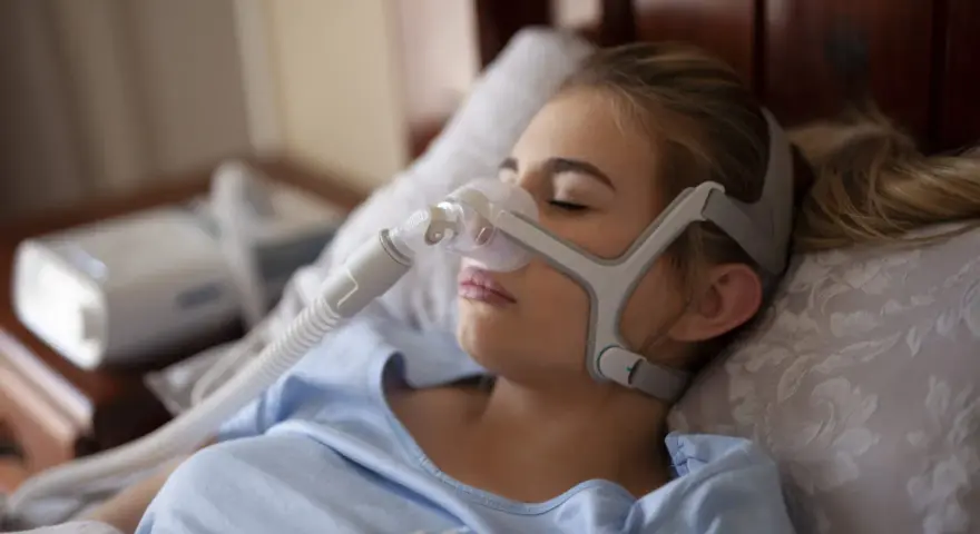 Kobieta śpi w masce CPAP, która pomaga w leczeniu bezdechu sennego. Urządzenie zapewnia ciągłe doprowadzanie powietrza, łagodząc objawy.