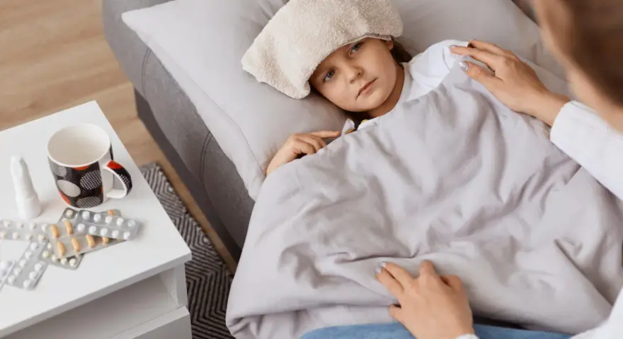 Chora dziewczynka z okładem na głowie leży pod kocem. Obok leki i kubek. Mama czuwa, zapewniając opiekę na dziecko.
