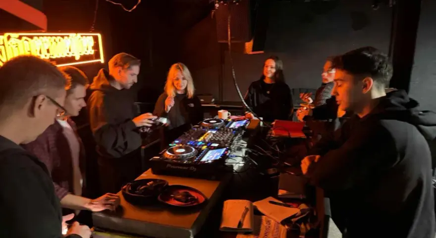Grupa młodych ludzi przy konsoli DJ-skiej, uczących się, jak zostać DJ-em. Neonowy napis w tle.