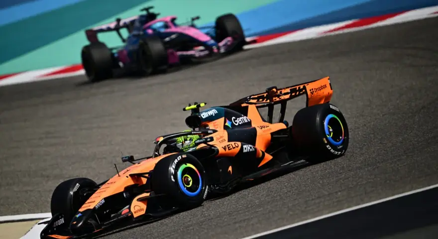 Un coche de Fórmula 1 naranja y negro compite en la pista, anticipando el reglamento F1 2026. Otro coche rosa y azul le sigue de cerca.