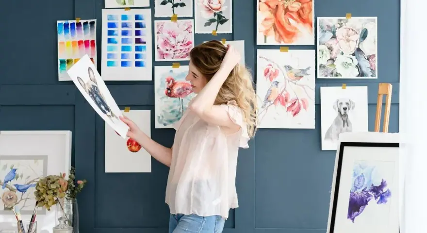 Mujer joven sonríe mientras revisa un dibujo de perro, rodeada de acuarelas de flores, aves y un flamenco. Un espacio inspirador para estudiar dibujo.