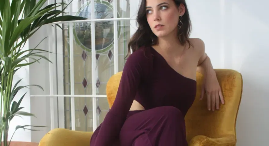 Mujer con escotes asimétricos en un sillón amarillo, junto a una planta y una ventana con vidrieras.