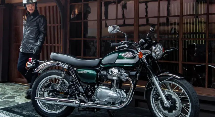 Mężczyzna w skórzanej kurtce i kasku stoi obok klasycznego motocykla Kawasaki W800 w kolorze ciemnozielonym.