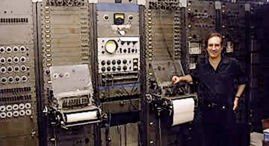 Mann steht neben einem riesigen, frühen Computer, dem ersten Synthesizer der Musikgeschichte.