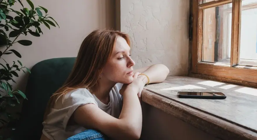 Zamyślona rudowłosa dziewczyna siedzi przy oknie, zastanawiając się, **benching co to** znaczy. Obok leży telefon.