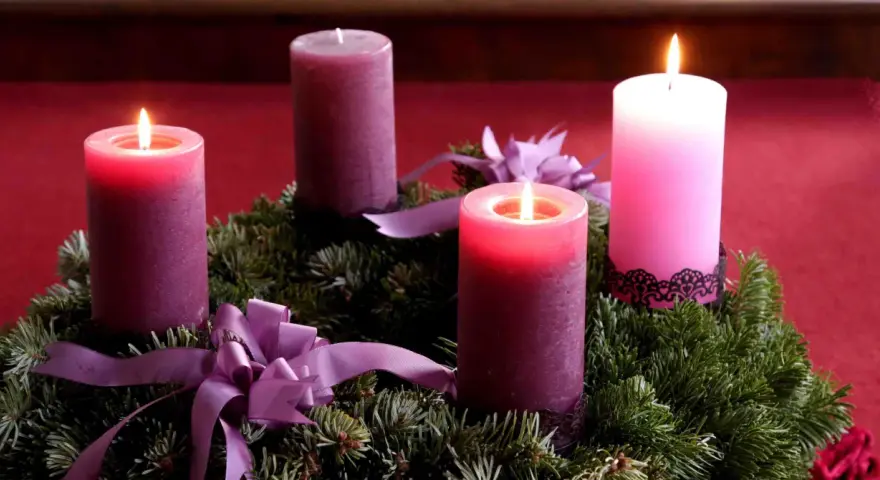 Drei brennende Kerzen auf einem Adventskranz aus Tannengrün mit lila Schleifen. Die dritte Kerze leuchtet hell.