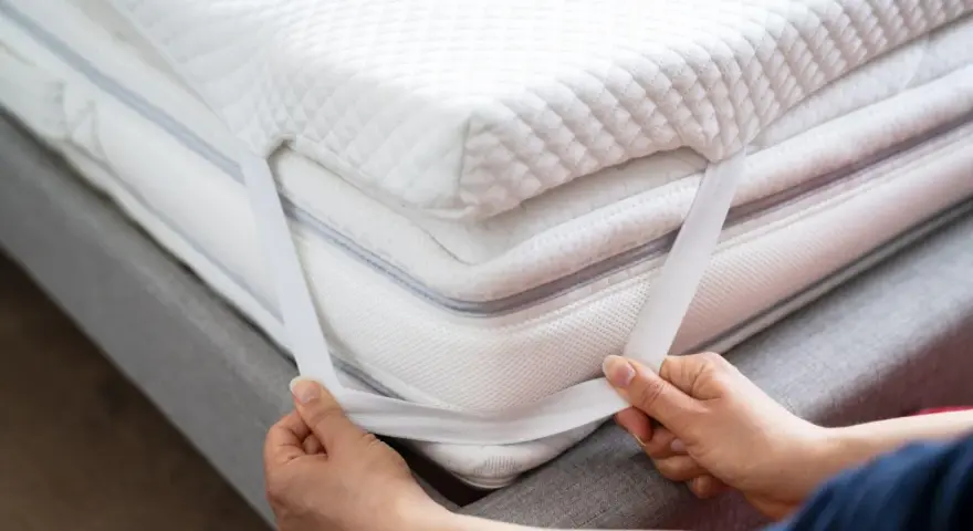 Mains ajustant les élastiques d'un surmatelas blanc et moelleux. Un surmatelas améliore le confort et la protection de votre matelas.