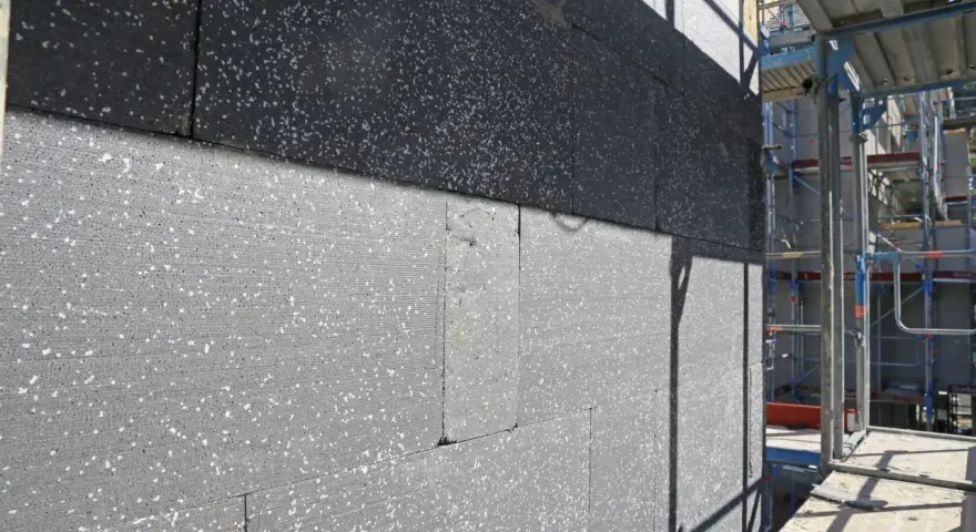 Gros plan sur des blocs de polystyrène gris, certains recouverts d'une couche noire, en cours d'installation pour une isolation par l'extérieur. Échafaudages visibles à droite.