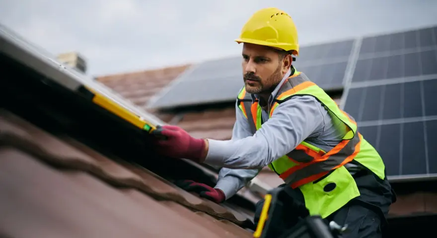 Instalator w żółtym kasku i kamizelce bezpieczeństwa montuje panele fotowoltaiczne na dachu. To dowód, że fotowoltaika jest bezpieczna, gdy montują ją profesjonaliści.