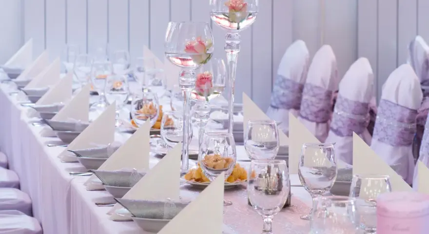 Vacker dukning med rosa rosor i glasvaser, vita servetter och silverfärgade skålar på en rosa bordsduk.