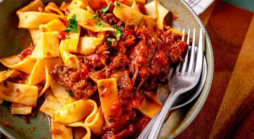 Breite Bandnudeln mit herzhaftem Ragu-Sauce, garniert mit frischer Petersilie. Ein Gabel liegt bereit.
