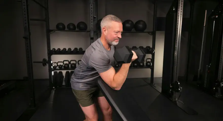 Man med grått hår och skägg utför preacher curls med en hantel i ett gym.