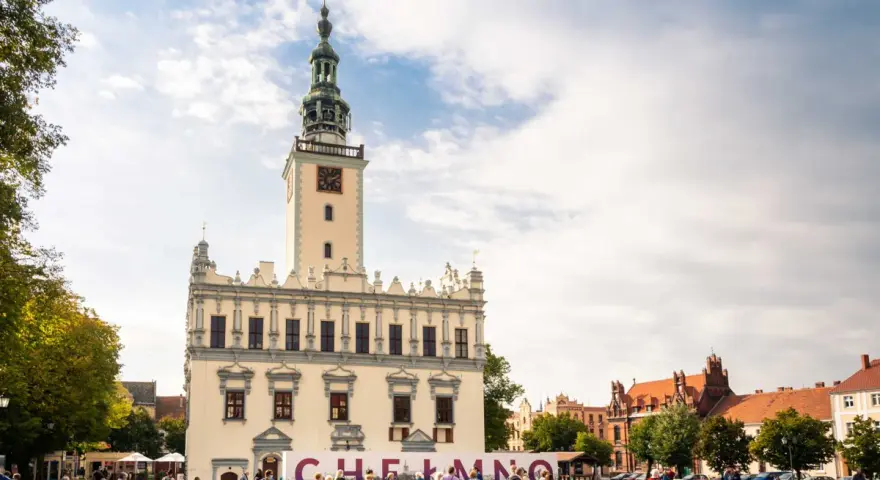 Rynek w Chełmnie z ratuszem i tablicą z napisem "Chełmno". Ludzie spacerują, podziwiając atrakcje miasta.
