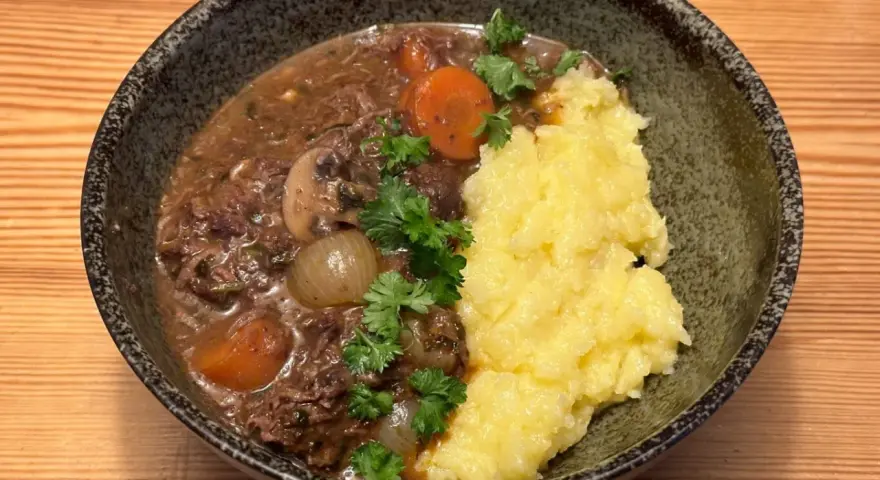 En skål med mustig biff bourguignon med möra köttbitar, champinjoner, lök och morötter, serverad med krämigt potatismos och garnerad med persilja.