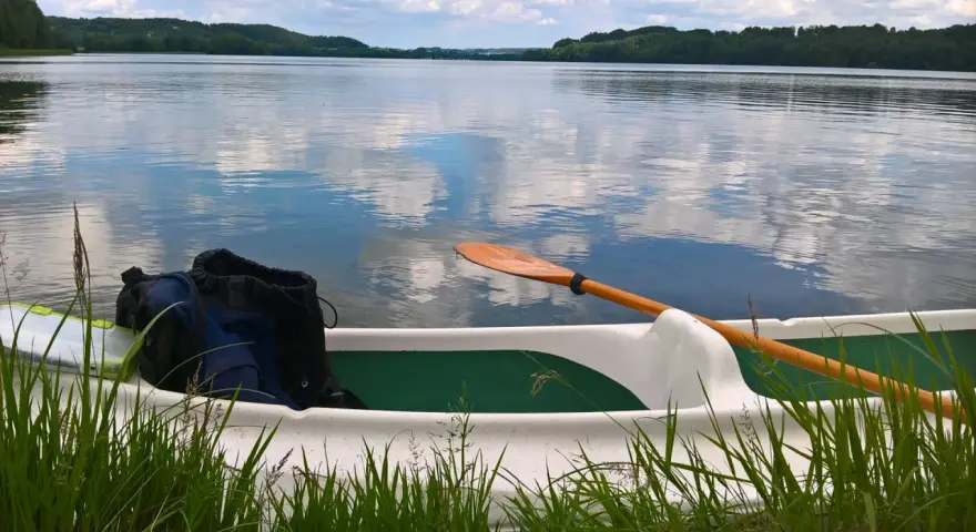 Wiosło i plecak w łodzi na spokojnym jeziorze raduńskim dolnym. Chmury odbijają się w tafli wody.