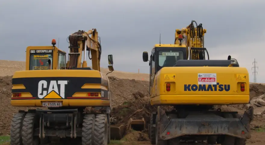 Dwie żółte koparki, CAT i Komatsu, stoją na placu budowy. Czy koparka ma dowód rejestracyjny? Na tablicy rejestracyjnej CAT widnieje KL 505 AE.