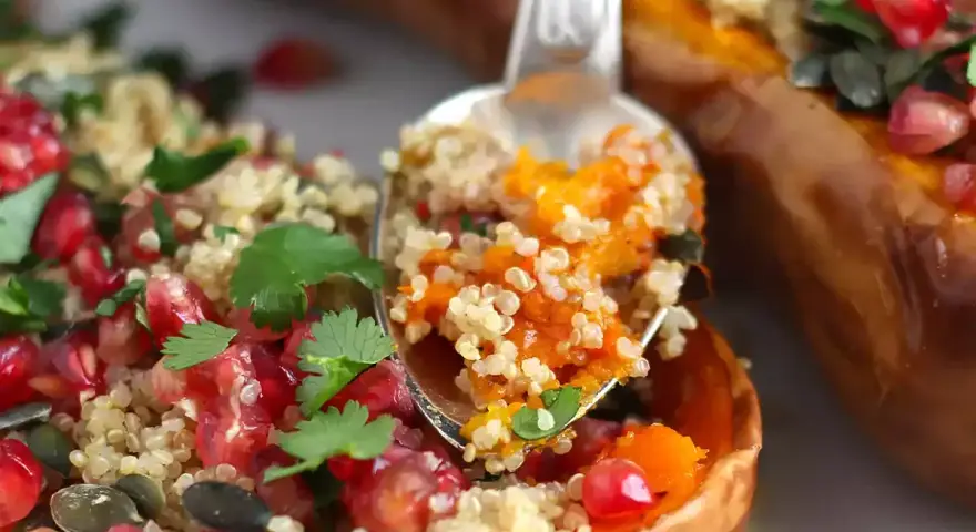 En sked tar en portion av ett läckert quinoa recept fyllt med pumpa, granatäpple och örter, serverat i en ugnsbakad butternut squash.