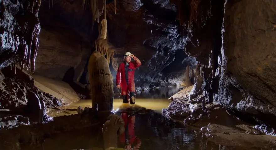 Eksplorator w jaskini, zanurzony po kolana w wodzie, oświetla drogę czołówką. To właśnie jest speleologia – odkrywanie podziemnego świata.