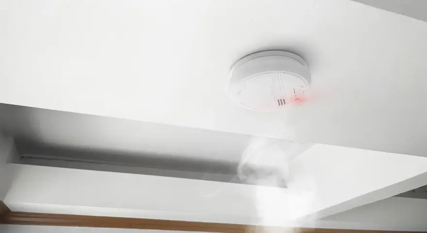 Detector de humo blanco en techo, emitiendo luz roja y humo. Cumpliendo la normativa de detectores de humo para seguridad.