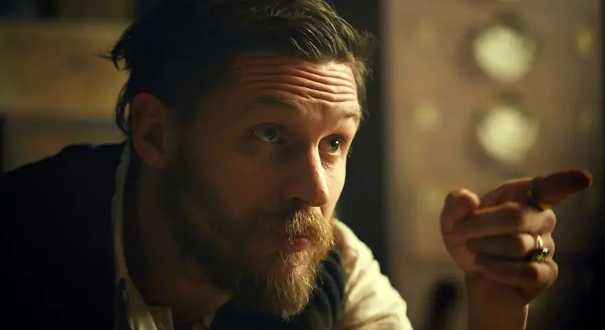 Mężczyzna z charakterystyczną fryzurą w stylu Peaky Blinders, z zarostem i pierścieniami na palcach, wskazuje palcem.
